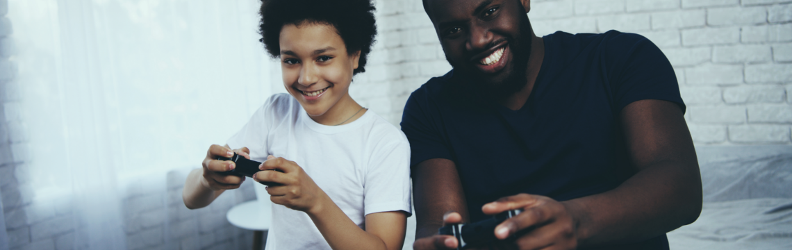 Young boy sitting next to his father, both holding video game cotnrollers, happily playing a video game