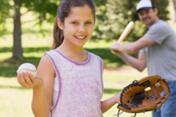 Tween girl holding a baseball with an adult male holding a baseball bat in the background