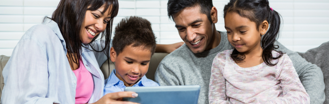 Family of four (two parents and two children) smile while holding and looking at a tablet computer